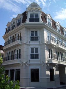 A four-story, cream-colored building with decorative balconies and a sign that reads AURUM on top, set against a partly cloudy blue sky. The architecture features large windows and ornate trim.