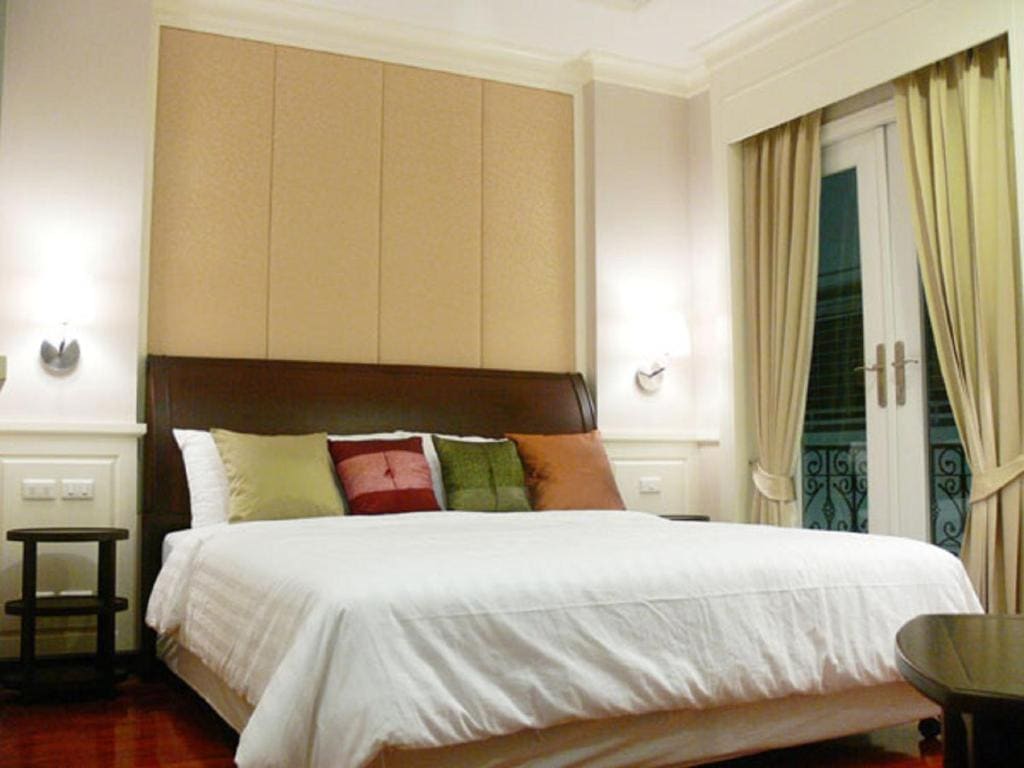 A neatly made bed with white linens and five colorful throw pillows sits in a modern bedroom with wall sconces, beige padded headboard, side tables, and large windows with cream curtains.
