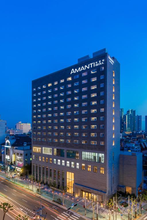 A tall, modern hotel building with the name AMANTI illuminated at the top, set along a busy city street at dusk with lights from passing cars and surrounding buildings visible.
