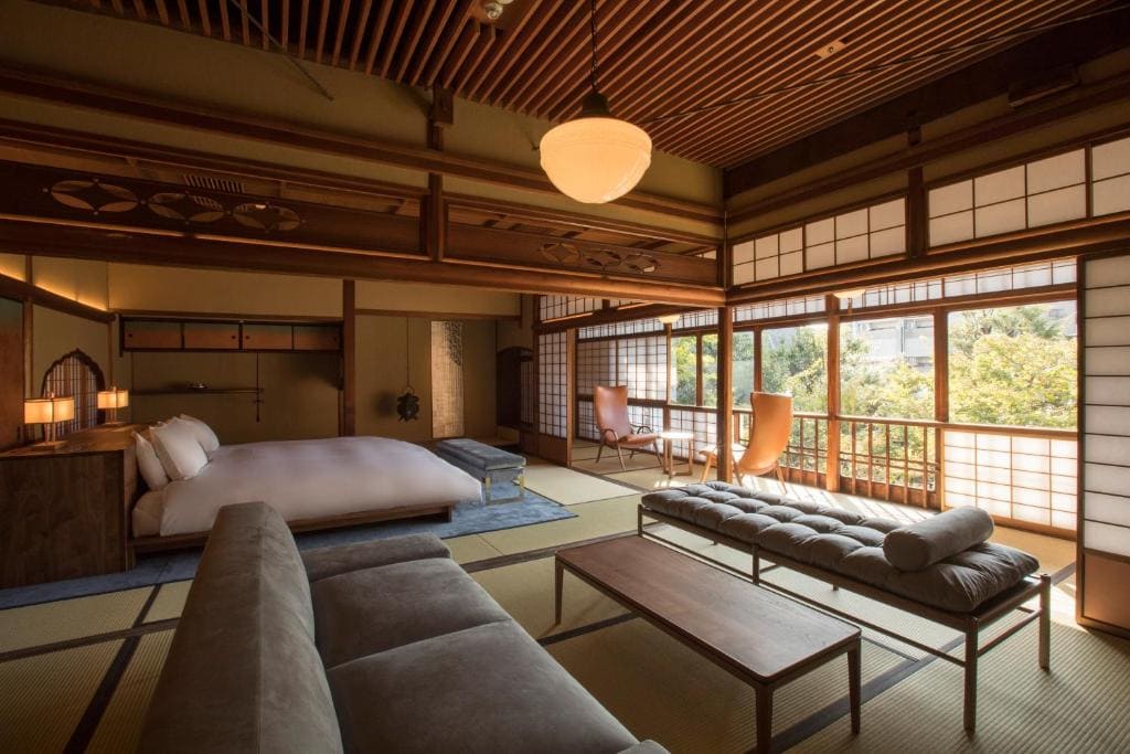 A spacious, elegant Japanese-style room with tatami mat flooring, a low bed, wooden furniture, large windows with shoji screens, and views of greenery outside, creating a serene and inviting atmosphere.