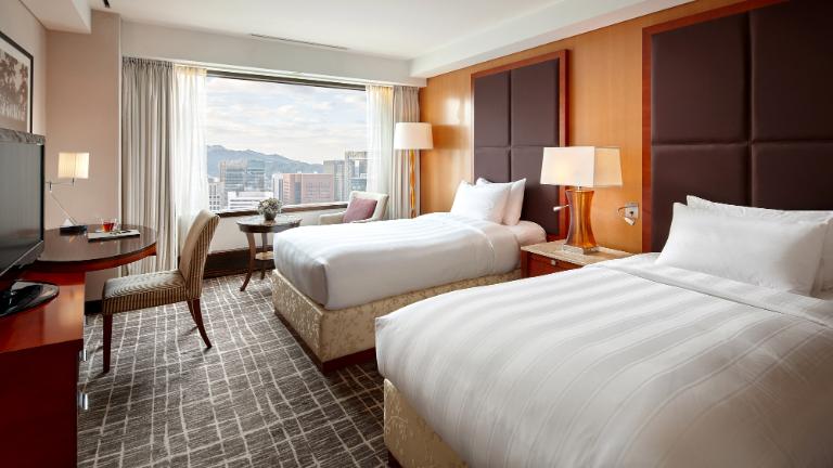 Modern hotel room with two twin beds, a large window showing a cityscape, a work desk with chair, lamps, and neutral-colored decor. Natural light brightens the room.