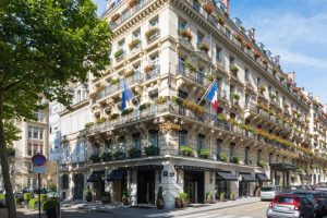 A grand, historic hotel building with ornate balconies adorned with flower boxes, French flags, and blue awnings on a sunny street corner, with trees, parked cars, and pedestrians nearby.