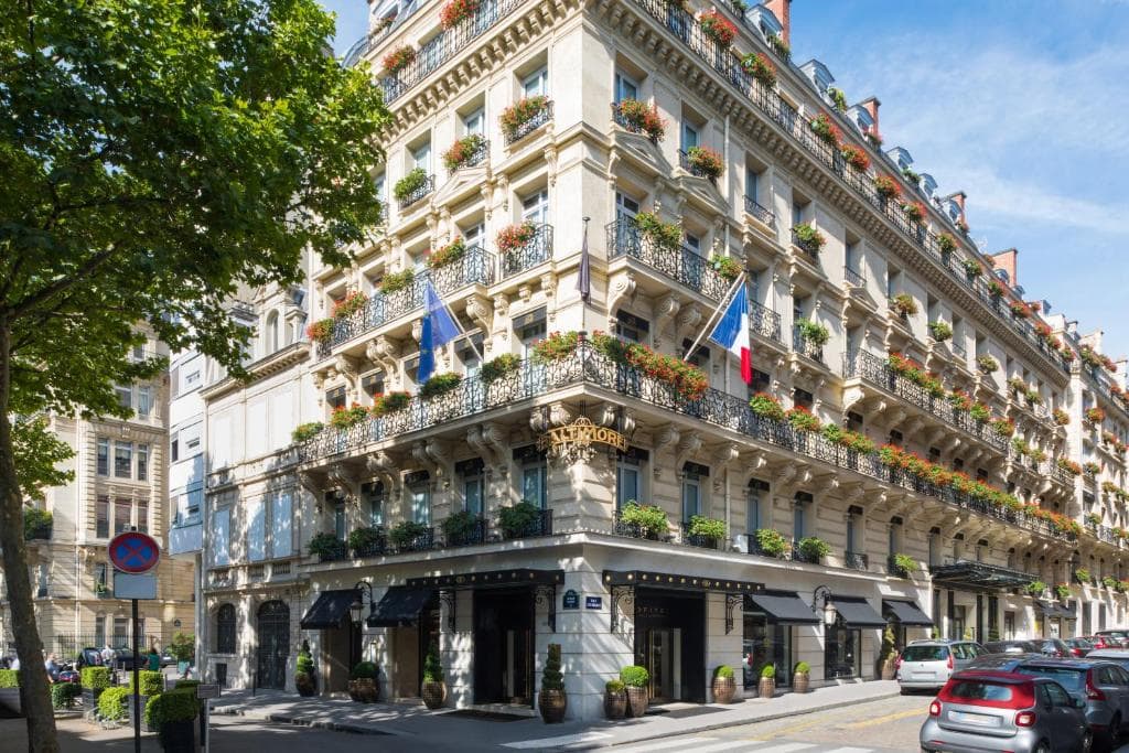 A grand, historic hotel building with ornate balconies adorned with flower boxes, French flags, and blue awnings on a sunny street corner, with trees, parked cars, and pedestrians nearby.