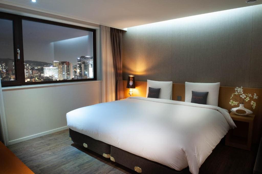 Modern hotel room with a large bed, two pillows, bedside lamps, wooden floor, and a window showing a cityscape at night with illuminated buildings. Warm lighting creates a cozy atmosphere.