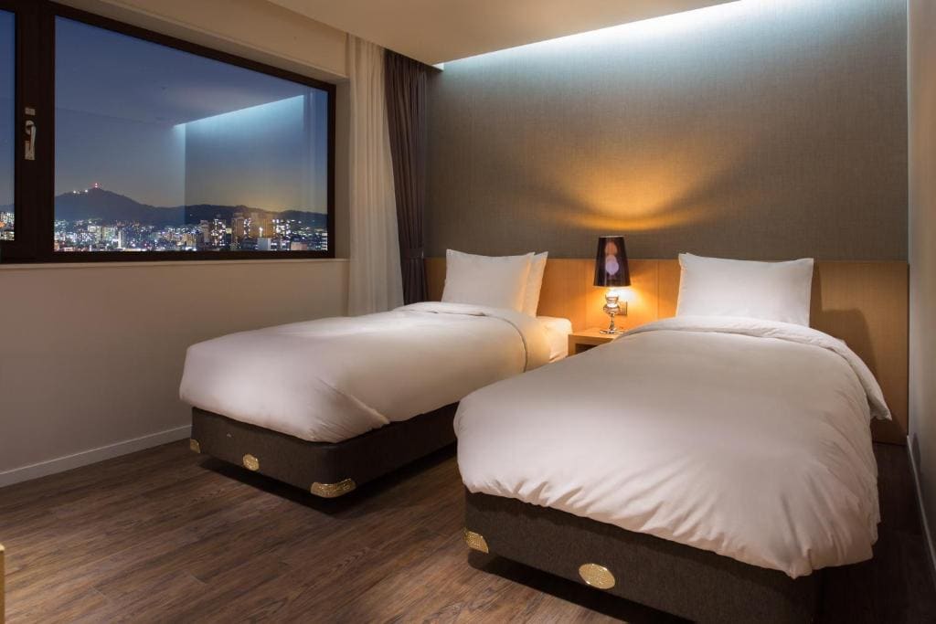 A modern hotel room with two neatly made single beds, a nightstand with a lamp and photo frame between them, wooden flooring, and a large window showing a cityscape at night.