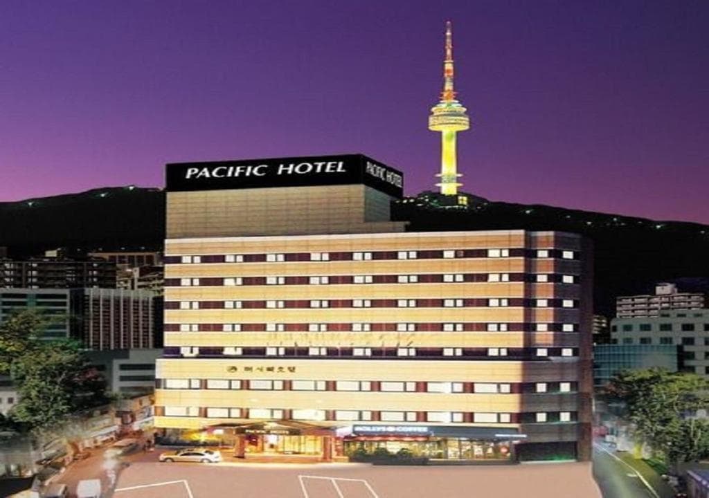 A multi-story building labeled Pacific Hotel is brightly lit at dusk, with the illuminated Namsan Seoul Tower visible on the hill behind, under a purple evening sky.