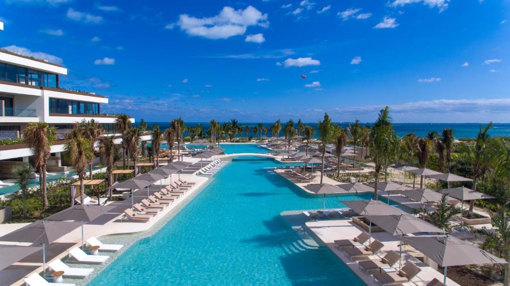 A luxury resort pool lined with lounge chairs and umbrellas, surrounded by palm trees, with buildings on the left and the ocean in the background under a bright blue sky.