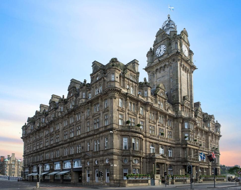 A grand, historic stone building with ornate architecture and a large clock tower, set against a blue sky. Flags are displayed near the entrance, and the street in front appears quiet and empty.