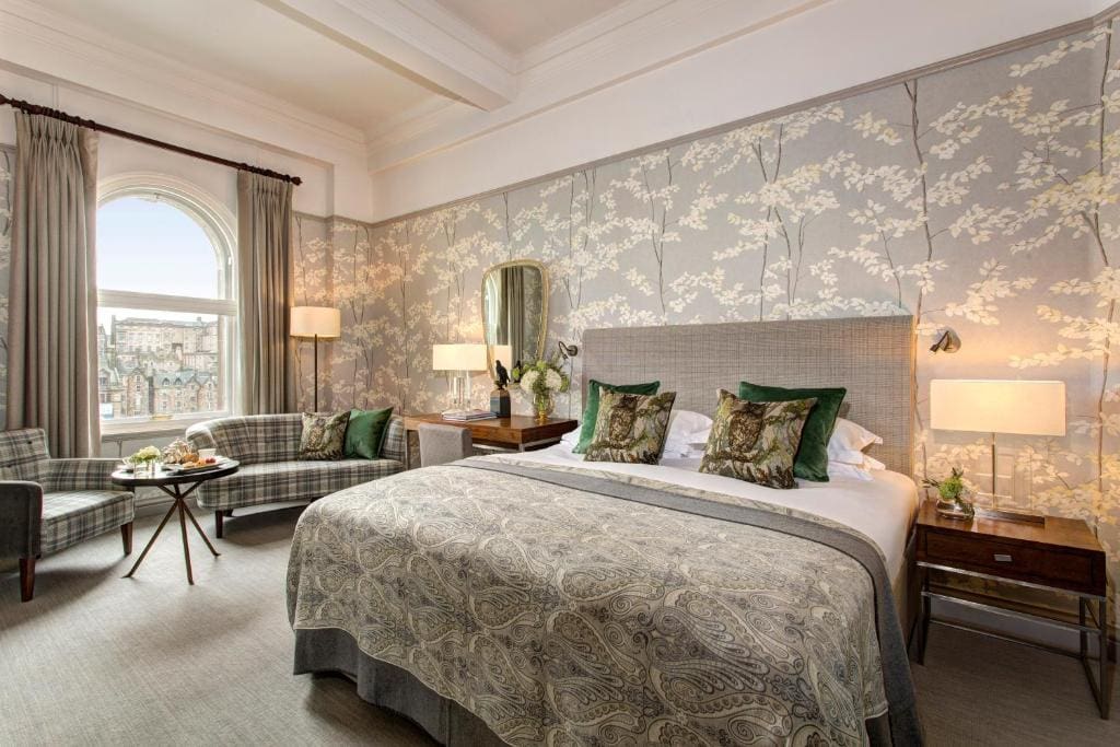 Elegant hotel room with a large bed, paisley bedspread, patterned wallpaper, plush pillows, a small sofa and chairs near a window with city views, bedside tables, lamps, and a decorative mirror.