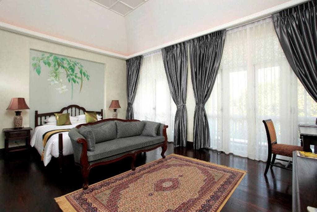 Bright bedroom with dark wood floors, a bed with white linens, a patterned rug, a classic sofa, two lamps, a desk with a chair, and large windows covered by sheer white curtains and dark drapes.