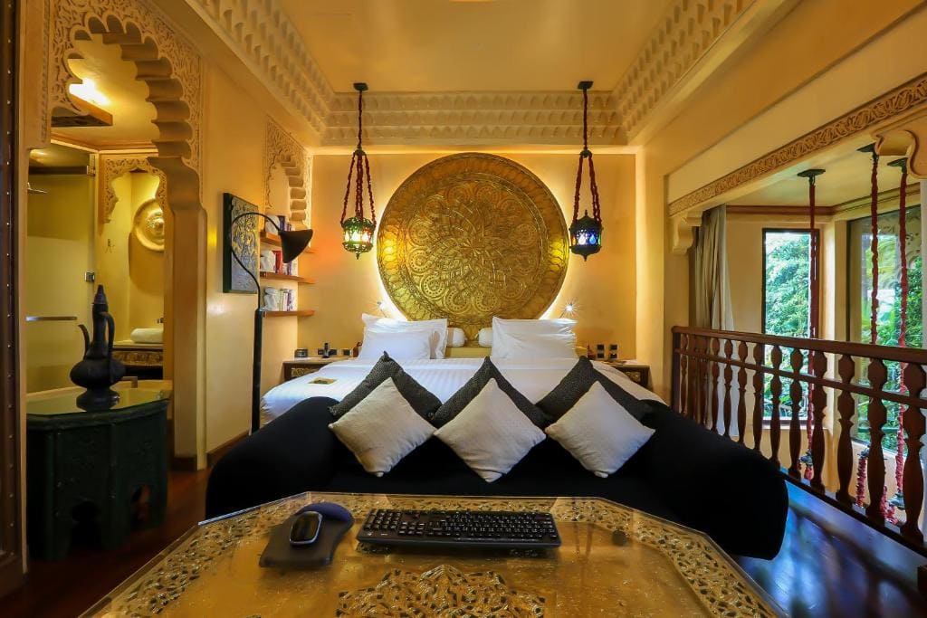 Luxurious bedroom with a large ornate golden medallion above the bed, stylish hanging lamps, black and white pillows, carved furniture, and large windows showing greenery outside.