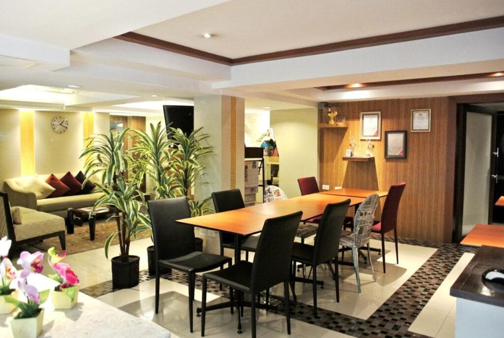 Modern, well-lit office lounge with a large wooden table and black chairs, potted plants, a cozy seating area with sofas in the background, and wall shelves displaying awards and framed certificates.