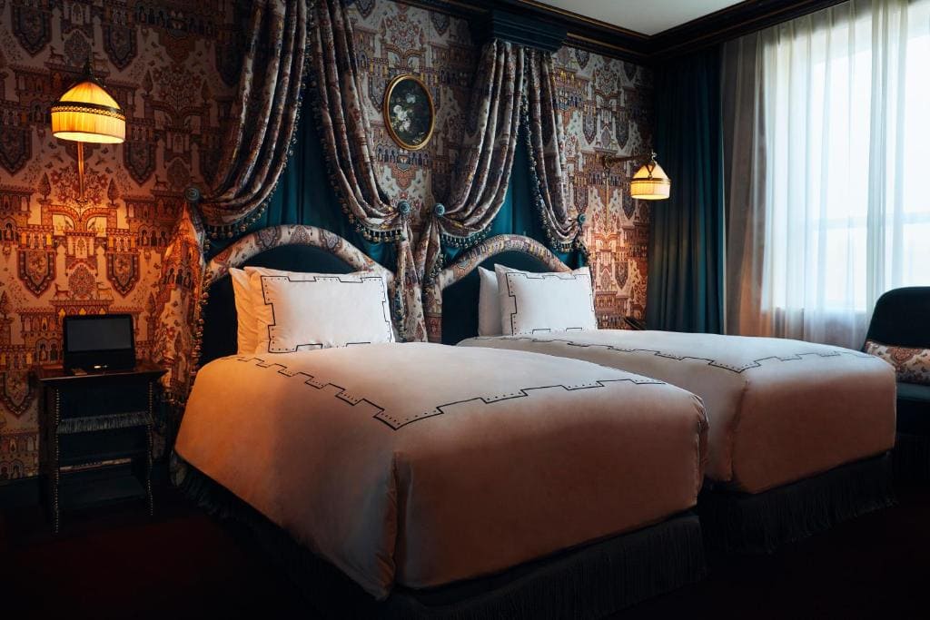 Twin beds with white linens in a vintage-style hotel room, featuring ornate wallpaper, draped fabric above the beds, two hanging lamps, a window with sheer curtains, and a framed oval portrait on the wall.