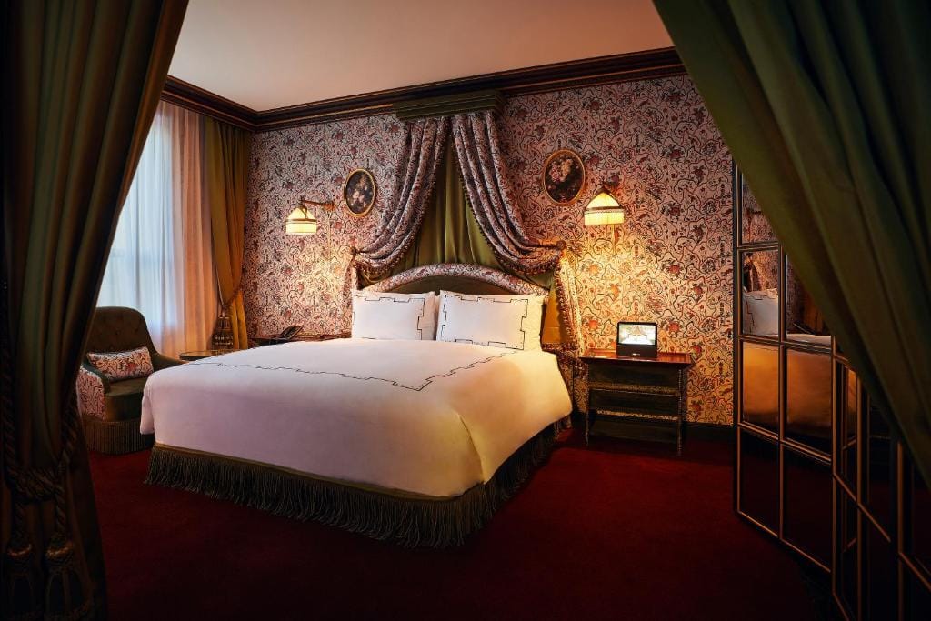 Elegant bedroom with a large bed, ornate headboard, and vintage wallpaper. Two framed portraits hang above, with warm lighting, heavy curtains, a plush chair by the window, and a mirrored wardrobe on the right.