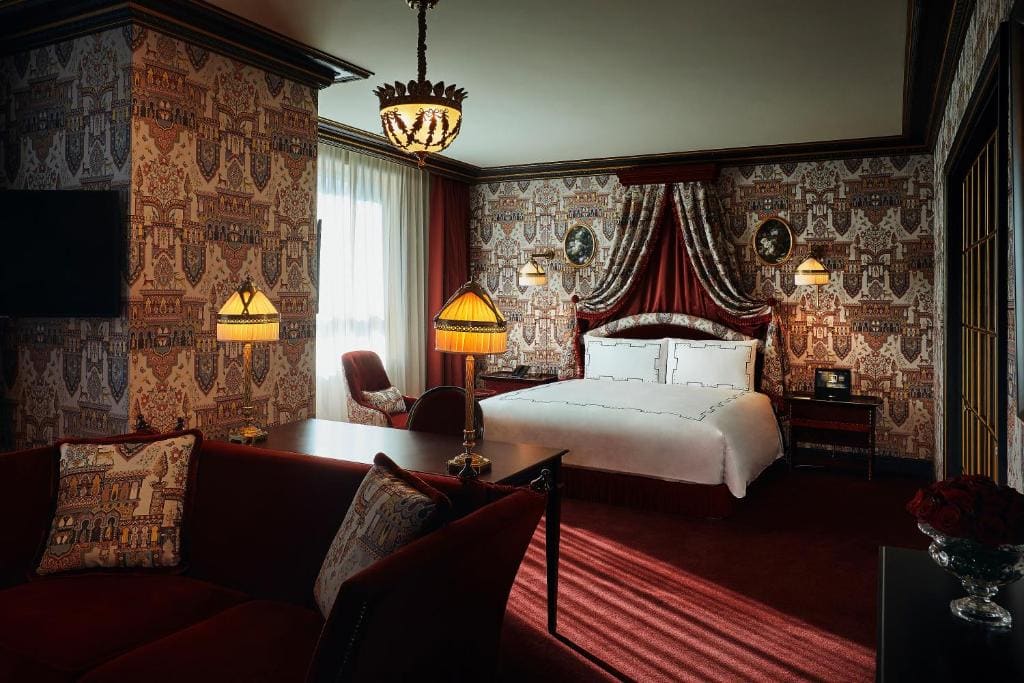 Luxurious hotel room with ornate wallpaper, elegant lighting, a velvet-upholstered bed with a canopy, deep red and gold accents, a sofa, armchair, and natural light coming through a window with sheer curtains.