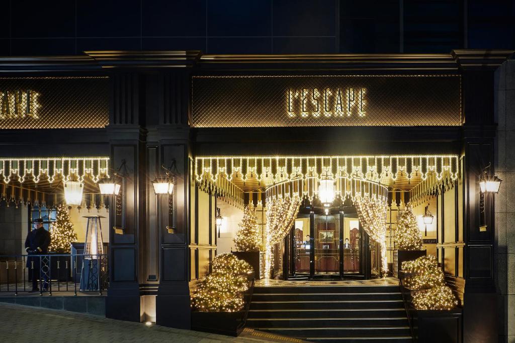 The entrance of a building decorated with warm white string lights and illuminated trees, with a sign above the door that reads LESCAPE. The overall ambiance is festive and inviting, suggesting a classy venue.