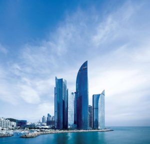 A cluster of modern glass skyscrapers stands by the waterfront under a partly cloudy blue sky, with reflections visible on the water and city buildings in the background.