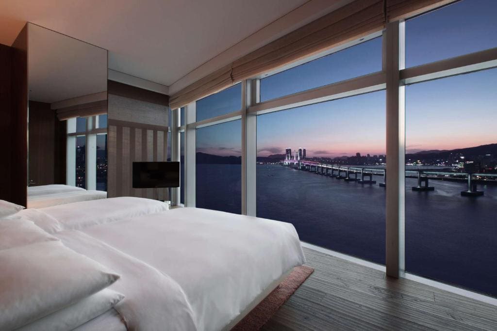 A modern hotel room with large floor-to-ceiling windows overlooking a calm body of water, a city skyline, and a bridge at dusk. The bed has white linens and there is a TV mounted on the wall.