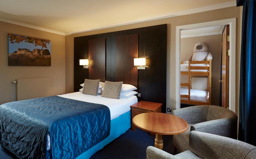 A hotel room with a double bed, two bedside tables, and two chairs with a small round table. In an adjacent room, there is a bunk bed with a large stuffed bear on the top bunk. The room has warm lighting.