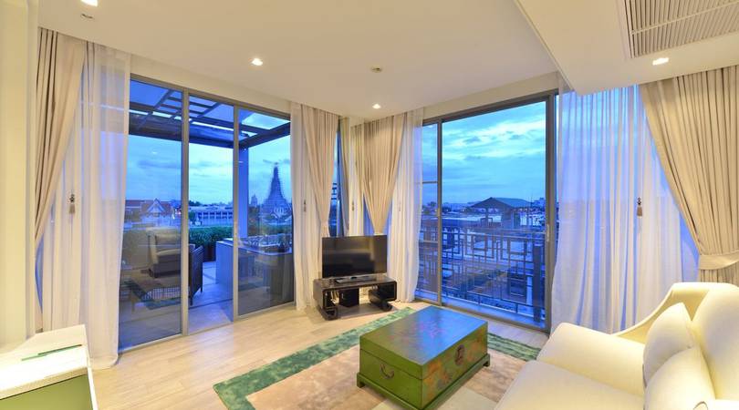 Modern living room with large floor-to-ceiling windows, sheer curtains, a small TV on a stand, a green coffee table, a white sofa, and a balcony with outdoor seating overlooking a cityscape at dusk.