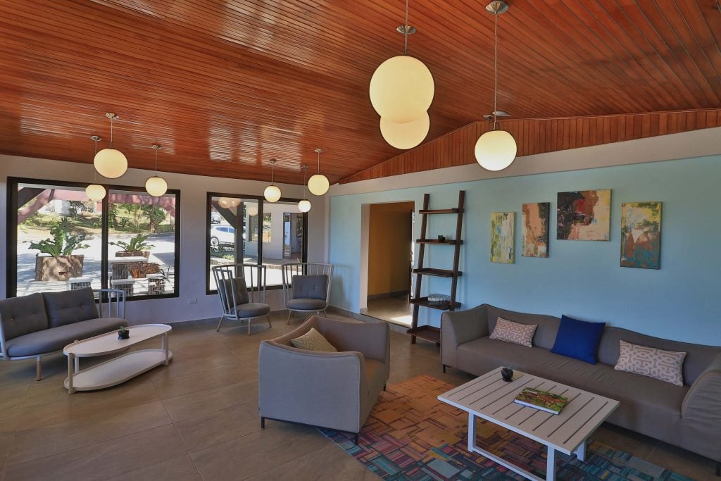 Modern lounge with wooden ceiling, round pendant lights, gray sofas and armchairs, a white coffee table, abstract wall art, and large windows overlooking greenery and parked cars outside.