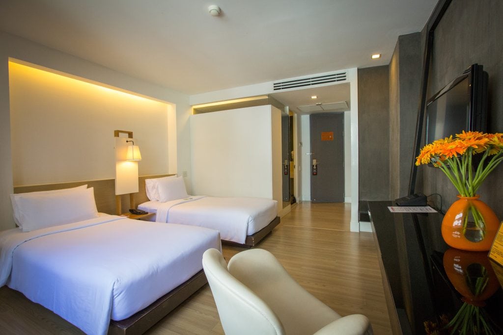 Modern hotel room with two neatly made single beds, a white chair, a flat-screen TV, wooden floor, and a vase of orange flowers on a dark desk. Soft lighting creates a warm, inviting atmosphere.