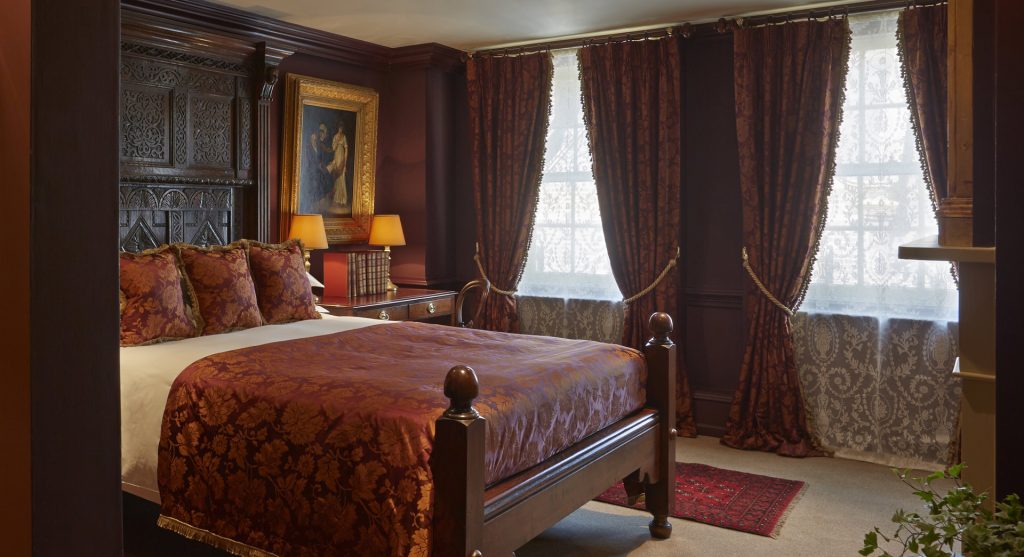 A richly decorated bedroom with dark wood furniture, ornate red and gold bedding, matching heavy drapes, lace-covered windows, a framed painting, and table lamps on either side of the bed.