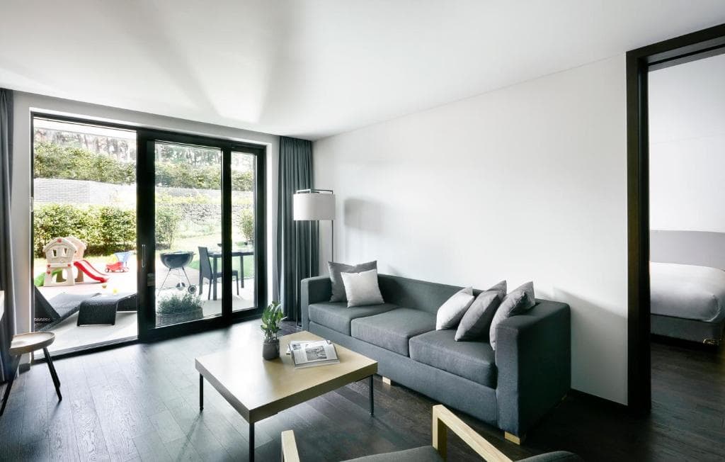 Modern living room with a gray sofa, coffee table, and floor lamp, opening onto a patio with outdoor furniture and a children’s playhouse; a bedroom is visible through an open door on the right.