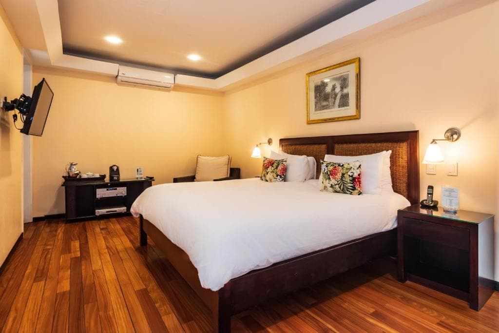 A cozy hotel room with a large bed, white bedding, floral pillows, bedside tables with lamps, wooden floor, a wall-mounted TV, air conditioner, and a coffee station in the corner. Framed art hangs above the bed.