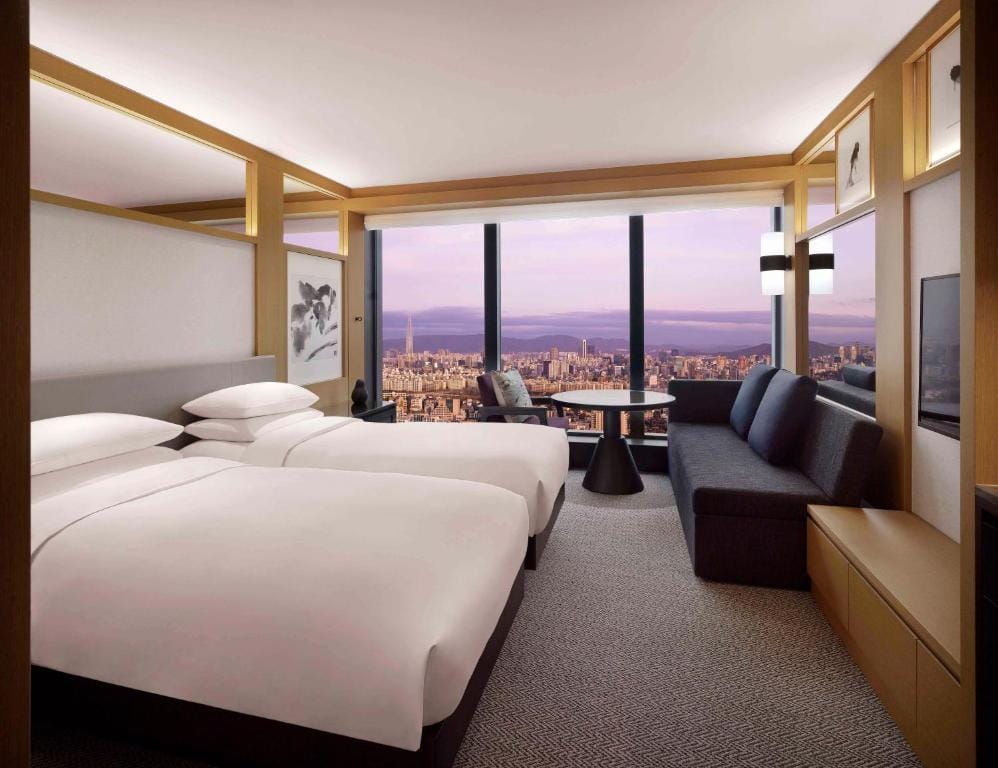 Modern hotel room with two large beds, a sofa, a table, and floor-to-ceiling windows offering a stunning cityscape view at sunset. Minimalist décor with warm wood accents and soft lighting.