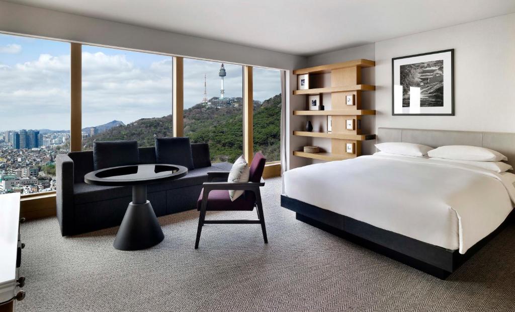 A modern hotel room with a large bed, sofa, table, chair, shelves, and a wide window offering a city view with hills, buildings, and a tall tower in the background. The room is bright and neatly arranged.