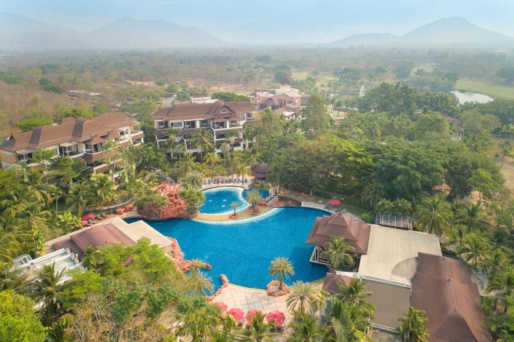 Aerial view of a large resort featuring a central outdoor swimming pool surrounded by palm trees, lush greenery, and several multi-story buildings, with hazy mountains visible in the background.