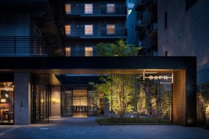 Modern hotel entrance at dusk with warm interior lighting, bamboo plants, and a glowing Bespoke sign. Tall apartments with illuminated windows rise in the background, creating an inviting urban atmosphere.