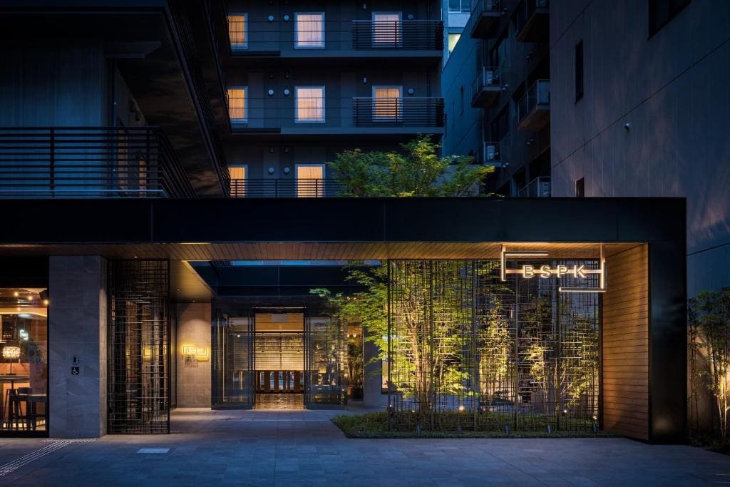 Modern hotel entrance at dusk with warm interior lighting, bamboo plants, and a glowing Bespoke sign. Tall apartments with illuminated windows rise in the background, creating an inviting urban atmosphere.