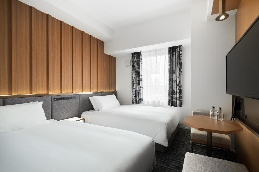 A modern hotel room with two double beds, white bedding, a wooden accent wall, a window with black-and-white curtains, a TV mounted on the wall, and a small table with two water bottles and a cushioned bench.