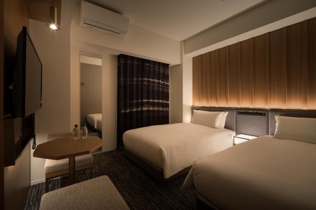 A modern hotel room with two twin beds, a wall-mounted TV, a small table with two water bottles, a mirror, and striped dark curtains. Warm lighting creates a cozy atmosphere.