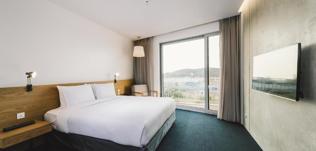 A modern hotel room with a large bed, wooden headboard, bedside tables, wall lamps, a chair by the window, floor-to-ceiling curtains, a flat-screen TV, and a view of a city and hills outside.