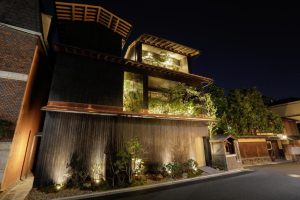 A modern three-story building with dark wooden exterior, illuminated at night. Plants and greenery are visible on balconies and in front, giving the structure a natural, serene appearance.