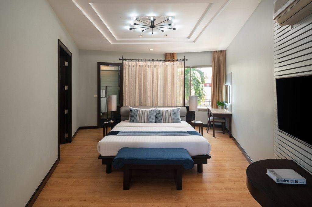 A modern hotel room with a large bed, blue bench, two bedside lamps, a desk by the window, wood flooring, neutral walls, and a contemporary ceiling light fixture.