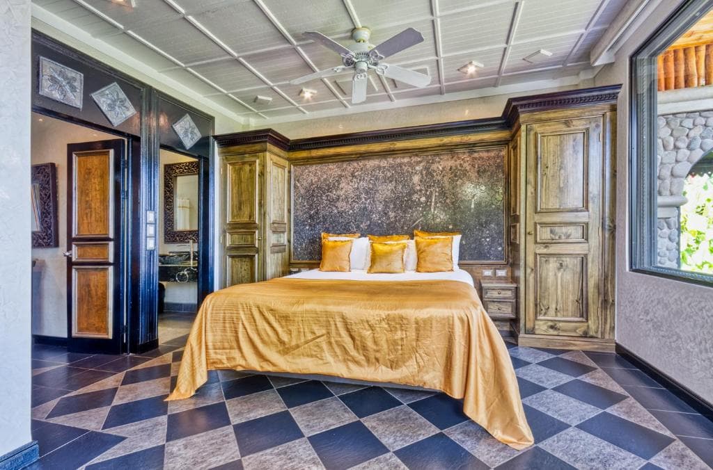Elegant bedroom with a double bed featuring gold bedding and pillows, flanked by wooden cabinets. The room has a checkered tile floor, a ceiling fan, and a window letting in natural light.