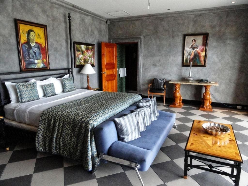 Elegant bedroom with a king-size bed, patterned bedding, a blue couch at the foot, checkered floor, wooden furniture, and three paintings on textured gray walls. A lamp and decor pieces add cozy touches.