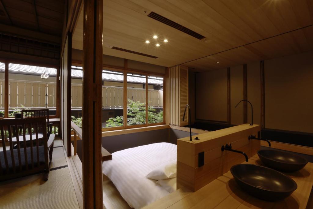 A modern Japanese-style room with a low bed, wooden furniture, two black vessel sinks, large windows overlooking a small garden, and a cozy balcony with chairs. Warm lighting enhances the serene, minimalist decor.