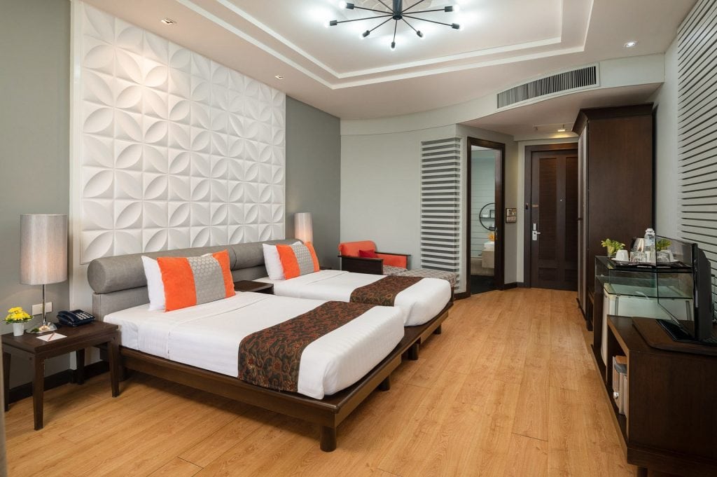 Modern hotel room with two single beds, white and orange accents, a geometric white accent wall, wooden flooring, a flat-screen TV, a desk, and a seating area near the door leading to a bathroom.