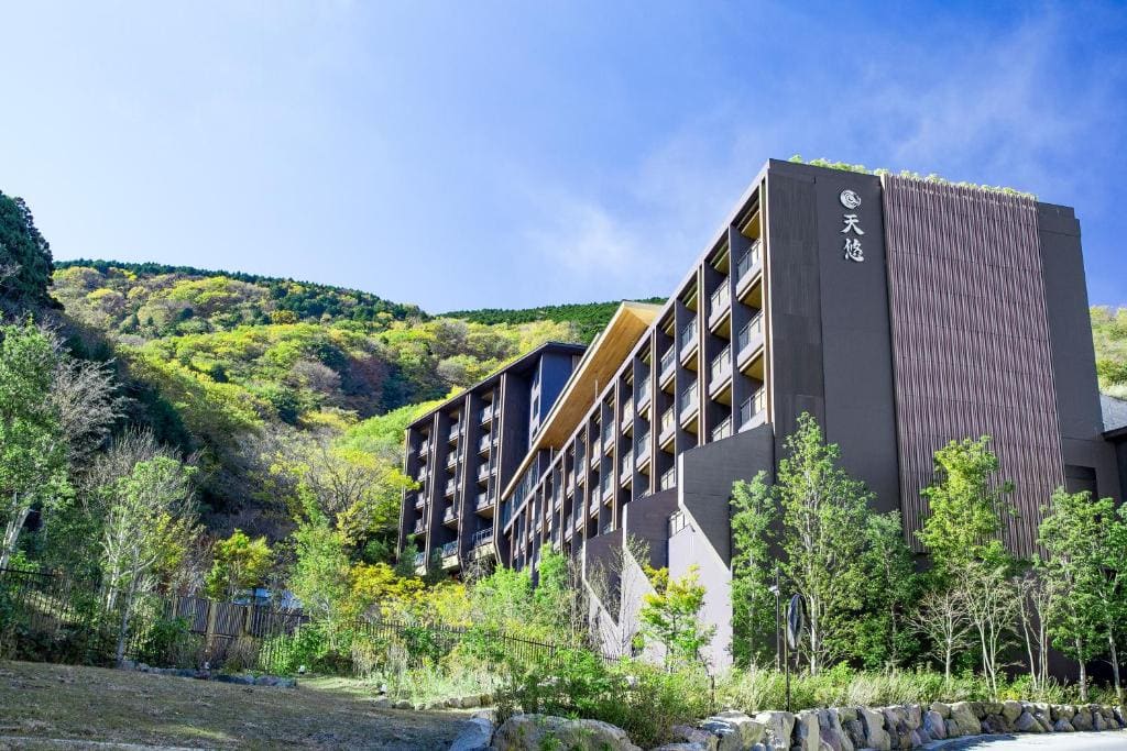 Modern multi-story hotel surrounded by lush greenery and trees, set against a forested hillside under a clear blue sky. Japanese characters are visible on the building’s facade.