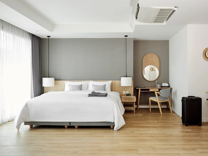 A modern hotel room with a large bed, two bedside tables, hanging lamps, a desk with a round mirror and chair, a suitcase, and wooden flooring. Natural light enters through sheer curtains.