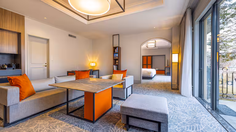 Modern hotel suite with a gray sofa, orange cushions, marble table, and ottoman in a spacious living area. Large windows let in natural light, and a bedroom is visible through an open archway.