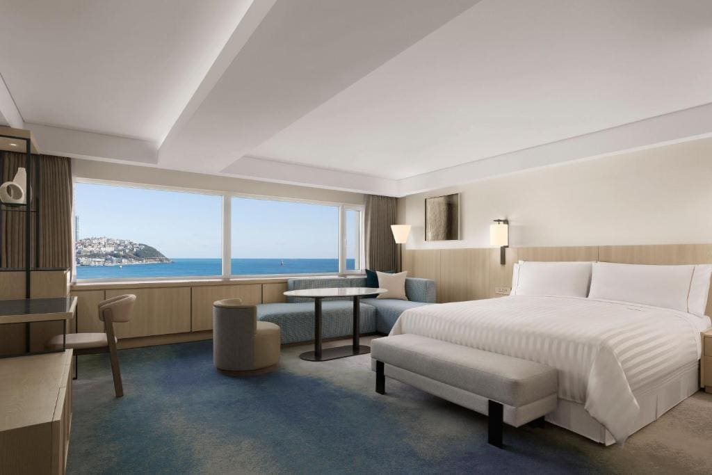 A spacious hotel room with a large bed, desk, and seating area, featuring a wide window with a view of the sea and a distant coastline under a clear sky.