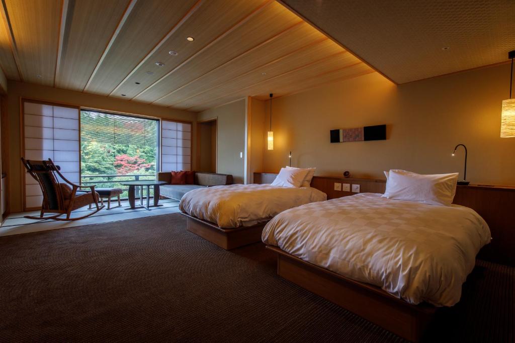 A spacious Japanese-style hotel room with two beds, a wooden ceiling, and large windows overlooking a garden with red and green trees. There is a rocking chair and a small table near the window.