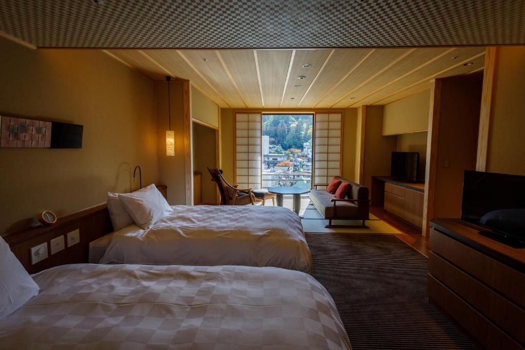 A cozy hotel room with two double beds, a TV, and a sitting area by large windows. The windows offer a scenic view of a town and hills outside. The room features warm lighting and traditional Japanese-style décor.