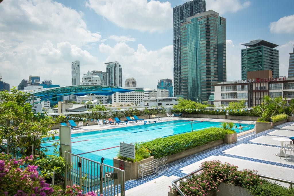 A rooftop swimming pool surrounded by greenery and lounge chairs, with tall modern skyscrapers and a partly cloudy blue sky in the background.